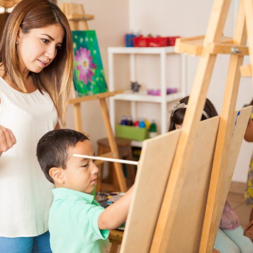 Pretty Hispanic art teacher reviewing and grading the work of a little boy during class